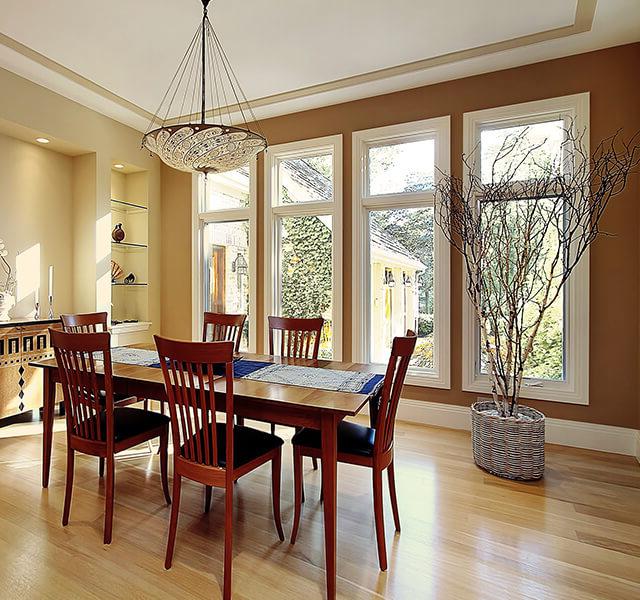 A dining room with a wooden table and six matching chairs. Large windows let in natural light, and a modern decorative chandelier hangs above. A potted tree stands in the corner next to built-in shelves displaying small items.